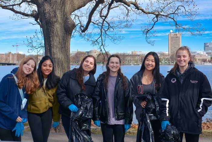 6 sorority members hold trash bags and pose for a photo next to the Charles river, with the Green buiding in the background