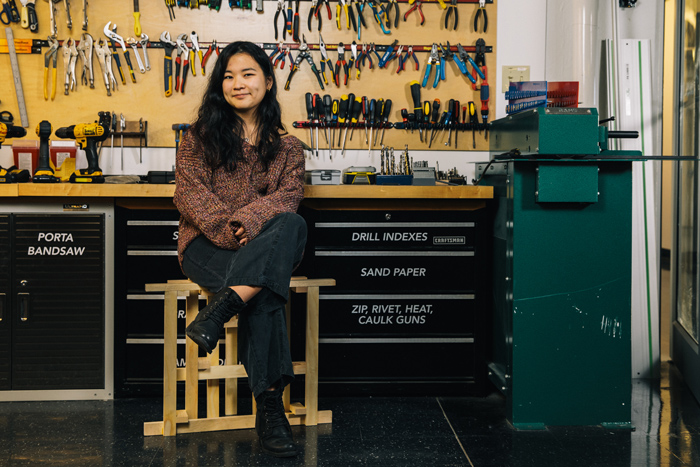 Photo of Ibuki Iwasaki sitting down in workshop with wall of tools in background