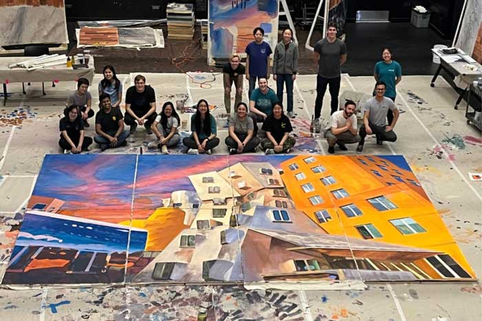 17 artists stand next to a gigantic painting of Stata center in oranges and blues.